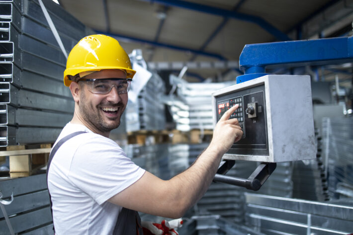 Trabalhador operando máquina industrial e ajustando parâmetros no computador para gerar renda extra em casa.